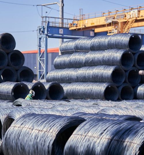 Large stacks of steel wire coils stored outdoors at an industrial site under bright daylight.